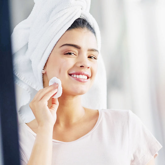 Woman applying cream to her face with a towel on her head, smiling.